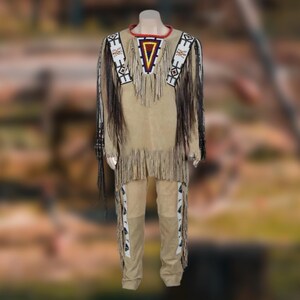 Op de afbeelding: Een bruin suède Native American outfit, bestaande uit een shirt en broek, tentoongesteld op een mannequin. Het shirt heeft een gefranjerde zoom, kralenaccenten en een geometrisch ontwerp bij de halslijn. De broek heeft decoratieve panelen langs de zijkanten.
