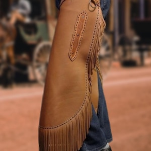 May include: Brown leather chaps with fringe detail, worn over blue jeans. The chaps have decorative stitching and a belt with a metal ring. The background is blurred, suggesting an outdoor setting.