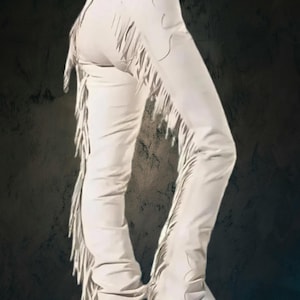Puede incluir: Pantalones de cuero blancos con cortes decorativos y largas flecos en los laterales. Los pantalones se muestran desde la cintura hacia abajo, con las piernas ligeramente dobladas. El fondo es una superficie oscura y texturizada.