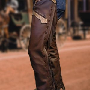 Puede incluir: Chaps de cuero marrón con costuras decorativas y flecos en los laterales. Los chaps tienen un acento de cuero marrón claro en la parte superior. Ropa de estilo western.