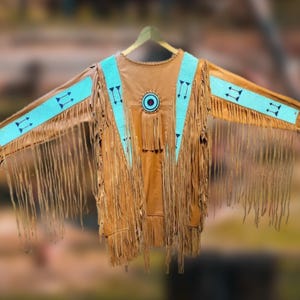Könnte beinhalten: Braune Lederjacke mit türkisfarbenen Akzenten und Fransen. Die Jacke hat ein rundes türkisfarbenes und schwarzes Design auf der Brust und türkisfarbene Bänder mit schwarzen Pfeildesigns auf den Ärmeln. Auf einem Holzbügel aufgehängt.