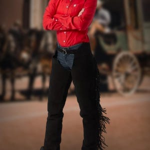 May include: A red long-sleeve button-down shirt paired with black suede chaps featuring fringe detailing. The outfit is completed with denim jeans and cowboy boots, creating a Western-inspired look.