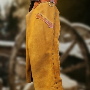 May include: A pair of tan suede leather chaps with brown leather accents. The chaps feature decorative stitching, fringe along the side, and a belt with a buckle. A metal ring is attached near the top.
