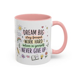 Può includere: Una tazza in ceramica bianca con manico e bordo rosa, che riporta il testo DREAM BIG, stay focused, WORK HARD, believe in yourself, NEVER GIVE UP. Il design include icone colorate disegnate a mano come un sole, un arcobaleno, una matita, un libro e una lampadina.