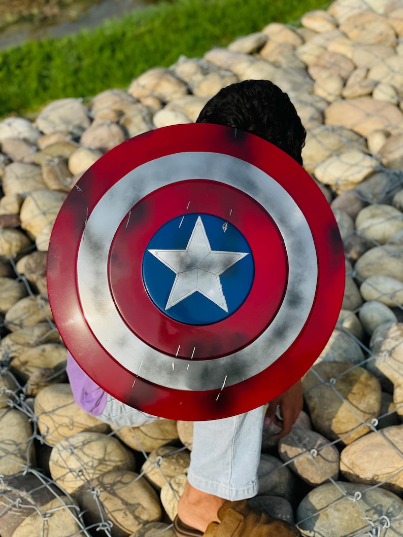K&ouml;nnte beinhalten: Ein Captain America Schild mit einem rot, wei&szlig; und blau Farbschema. Der Schild hat einen zentralen blauen Kreis mit einem wei&szlig;en Stern, umgeben von konzentrischen roten und wei&szlig;en Ringen. Der runde Schild scheint aus Metall zu sein.