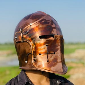 May include: A copper-coloured medieval helmet with a cross design on the face guard. The helmet has a polished finish and a chain detail on the side. The background is a blurred view of a field and sky.