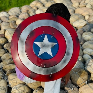 K&ouml;nnte beinhalten: Ein Captain America Schild mit einem rot, wei&szlig; und blau Farbschema. Der Schild hat einen zentralen blauen Kreis mit einem wei&szlig;en Stern, umgeben von konzentrischen roten und wei&szlig;en Ringen. Der runde Schild scheint aus Metall zu sein.