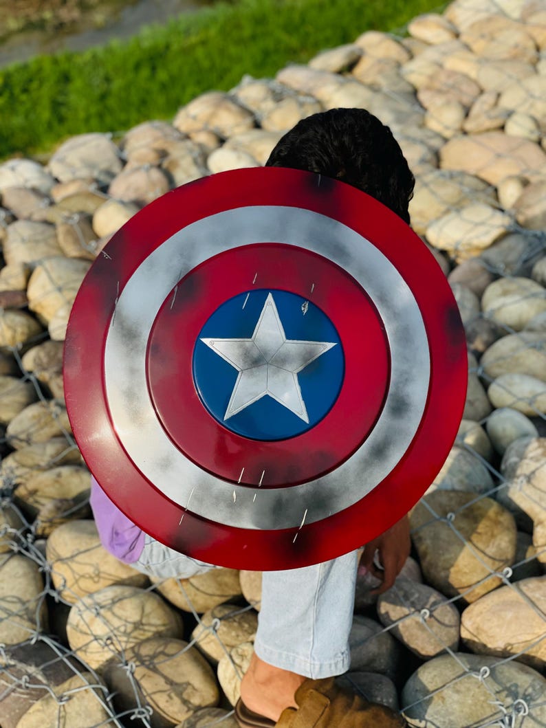 K&ouml;nnte beinhalten: Ein Captain America Schild mit einem rot, wei&szlig; und blau Farbschema. Der Schild hat einen zentralen blauen Kreis mit einem wei&szlig;en Stern, umgeben von konzentrischen roten und wei&szlig;en Ringen. Der runde Schild scheint aus Metall zu sein.