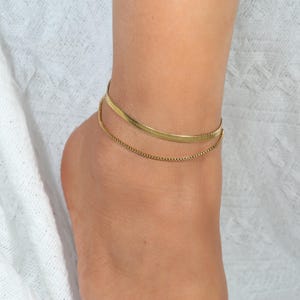 Op de afbeelding: Een goudkleurige, dubbellaagse enkelband. De bovenste laag heeft een plat, getextureerd ontwerp, terwijl de onderste laag een delicate ketting is. De enkelband wordt op de enkel gedragen.