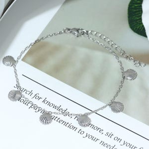 May include: A silver-colored anklet featuring small seashell charms. The delicate chain has an adjustable length and a lobster clasp closure. The anklet is displayed on a white surface with text that reads "Search for knowledge, remore, sit on your hout payi attention to your needs."
