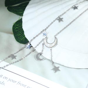 Puede incluir: Un collar plateado en capas con colgantes de estrella y media luna. El collar tiene dos hilos, uno con pequeños colgantes de estrella y otro con un colgante de luna más grande y estrellas colgantes. El colgante de luna tiene una incrustación de nácar.