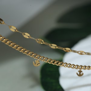 Może przedstawiać: Zbliżenie dwóch złotych naszyjników. Górny naszyjnik ma delikatny łańcuszek z owalnych ogniw. Dolny naszyjnik posiada łańcuch typu curb z małymi zawieszkami w kształcie kotwicy. Biżuteria jest prezentowana na rozmytym tle.