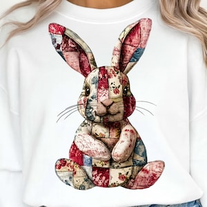 Peut inclure: Sweat-shirt blanc avec un motif de lapin patchwork. Le lapin est fait de tissu rouge, bleu et crème avec des motifs floraux. Le lapin est assis, les oreilles dressées, et des moustaches sont visibles.