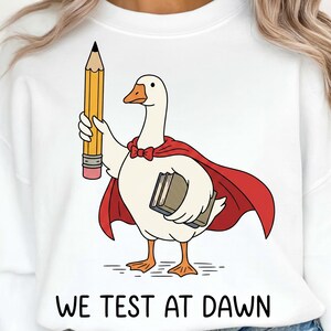 Puede incluir: Sudadera blanca con un ganso de dibujos animados con capa roja y pajarita, sosteniendo un gran lápiz amarillo y un libro. El texto "WE TEST AT DAWN" está impreso en negro en la parte inferior.