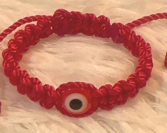 May include: Red woven bracelet featuring a central evil eye charm. The charm has a white iris, black pupil, and red surround. The bracelet is made of a thick, woven material and is adjustable.