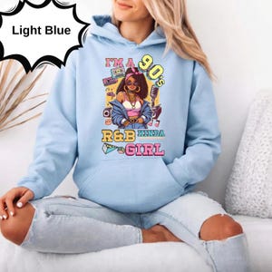 May include: Light blue hoodie with a graphic of a woman in 90s style, with the text "I'm a 90's R&B Kinda Girl". The graphic includes a boombox, microphone, and other 90s-themed elements. The hoodie is made of a soft material.