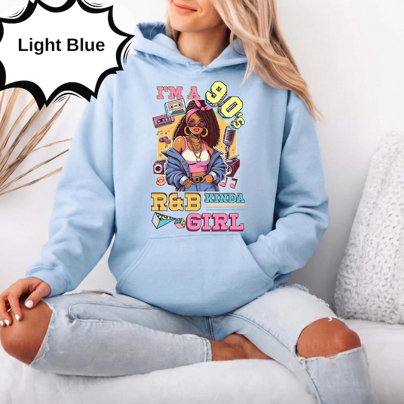 May include: Light blue hoodie with a graphic of a woman in 90s style, with the text "I'm a 90's R&B Kinda Girl". The graphic includes a boombox, microphone, and other 90s-themed elements. The hoodie is made of a soft material.