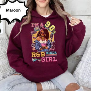 May include: Maroon hoodie with a graphic that reads "I'm a 90's R&B Kinda Girl." The graphic features a cartoon woman wearing sunglasses, a denim jacket, and a pink top. The design includes a cassette tape, microphone, and musical notes.