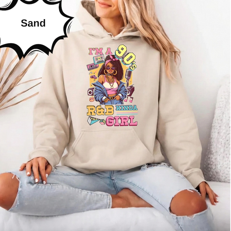 May include: Sand-colored hoodie with a graphic that reads "I'm a 90's R&B Kinda Girl." The graphic features a cartoon woman with sunglasses, a pink top, and a blue jacket. The hoodie has a drawstring hood and a kangaroo pocket.