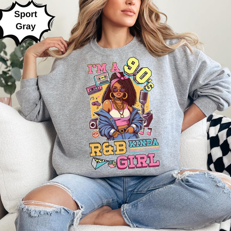 May include: Sport gray sweatshirt with a colorful graphic. The graphic features a woman with sunglasses and the text "I'm a 90's R&B Kinda Girl". The design includes a boombox, cassette tapes, and a microphone. The sweatshirt is a casual style.