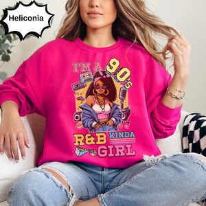 May include: A bright pink sweatshirt with a graphic featuring a 90s-themed illustration of a woman, a cassette tape, and a microphone. The text on the sweatshirt reads "I'm a 90's R&B Kinda Girl."