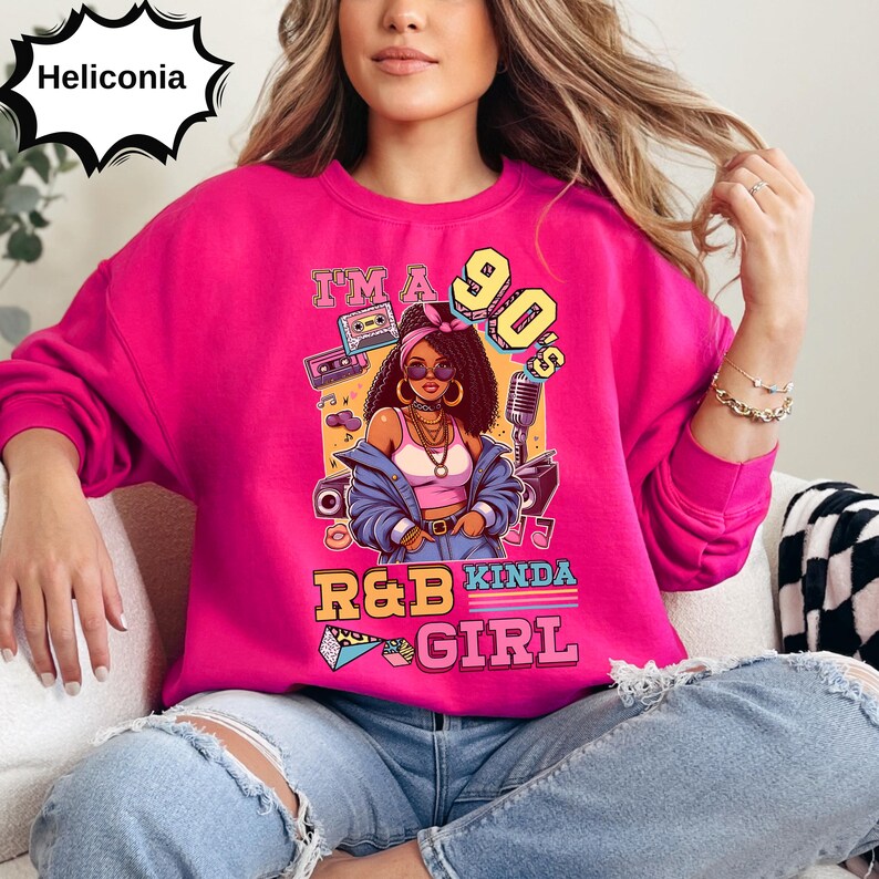 May include: A bright pink sweatshirt with a graphic featuring a 90s-themed illustration of a woman, a cassette tape, and a microphone. The text on the sweatshirt reads "I'm a 90's R&B Kinda Girl."