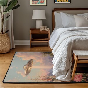 May include: A rectangular area rug featuring a surreal landscape with a golden UFO hovering above a pastel-colored sky. The rug has a black border and depicts mountains, flowers, and clouds. The rug is placed in a bedroom setting.