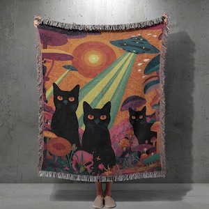 May include: A woven throw blanket featuring a colorful illustration of three black cats with orange eyes, mushrooms, and a UFO. The background has an orange and yellow sunset with green and yellow beams of light.