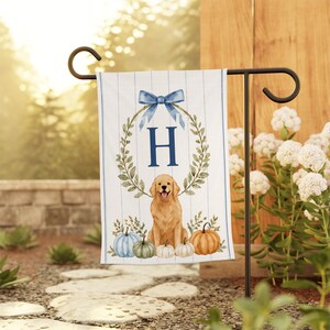 May include: A rectangular garden flag featuring a white background with thin blue vertical stripes. The design includes a central blue letter H inside a green leafy wreath topped with a blue bow, a golden retriever, and several pumpkins.
