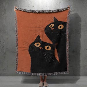 Könnte beinhalten: Eine gewebte Decke mit einer skurrilen Illustration von zwei schwarzen Katzen mit großen gelben Augen auf orangefarbenem Hintergrund. Die Decke hat einen Fransenrand und wird von einer Person gehalten.