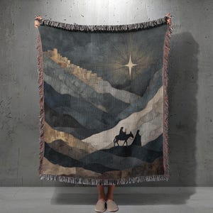 Könnte beinhalten: Eine gewebte Wurkdecke mit einer Landschaftsszene mit einem Stern, Bergen und Figuren auf einem Esel. Die Farbpalette umfasst Blau-, Grau- und Goldtöne. Die Decke hat einen Fransenrand.