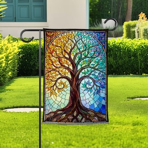 May include: A rectangular garden flag featuring a stained glass style design of a tree with a brown trunk and roots. The leaves transition from warm yellow and orange on the left side to cool green, teal, and blue tones on the right side.