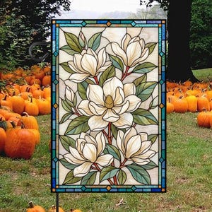 May include: A rectangular garden flag featuring a stained glass design of white magnolia flowers with green leaves. The border consists of blue and teal geometric glass segments. The artwork is displayed on a black metal stand.