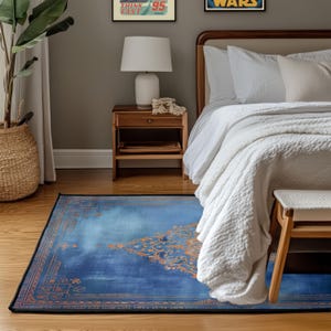 May include: A rectangular blue rug with an ornate orange design and a black border. The rug is placed on a wooden floor in a bedroom setting, with a bed, nightstand, and a woven basket visible in the background.