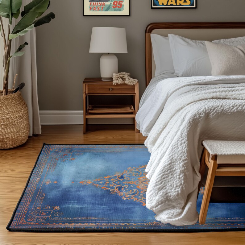 May include: A rectangular blue rug with an ornate orange design and a black border. The rug is placed on a wooden floor in a bedroom setting, with a bed, nightstand, and a woven basket visible in the background.