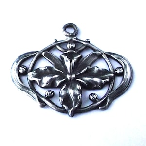 May include: A silver-toned pendant with an Art Nouveau floral design. The pendant features a central flower with four petals, surrounded by curved lines and small, round accents. It has a loop at the top for a chain.