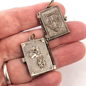May include: A silver book-shaped locket, open to show an embossed floral design. The closed side displays the letters "IHS" within a heart-shaped shield. The locket has a hinged spine and a small loop for a chain.