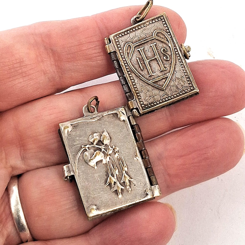 May include: A silver book-shaped locket, open to show an embossed floral design. The closed side displays the letters "IHS" within a heart-shaped shield. The locket has a hinged spine and a small loop for a chain.