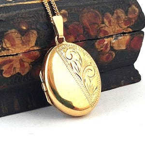 May include: A gold-colored oval locket pendant with an ornate floral design on a delicate gold chain. The locket is closed and has a shiny finish. The background is a dark wooden box with floral patterns.