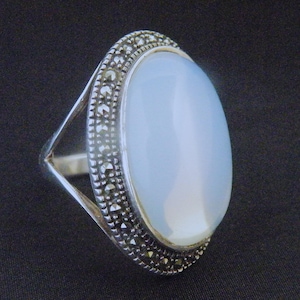May include: Oval-shaped silver ring with a large, pale blue gemstone. The gemstone is surrounded by a border of small, dark stones. The ring's band has a simple, open design. The ring is set against a dark background.