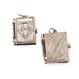 May include: Two small, silver-coloured book-shaped pendants. One pendant features the letters "IHS" within a heart-shaped design. The other pendant is open, displaying the word "LOURDES" with a wheat stalk design. Both pendants have a hinged design.