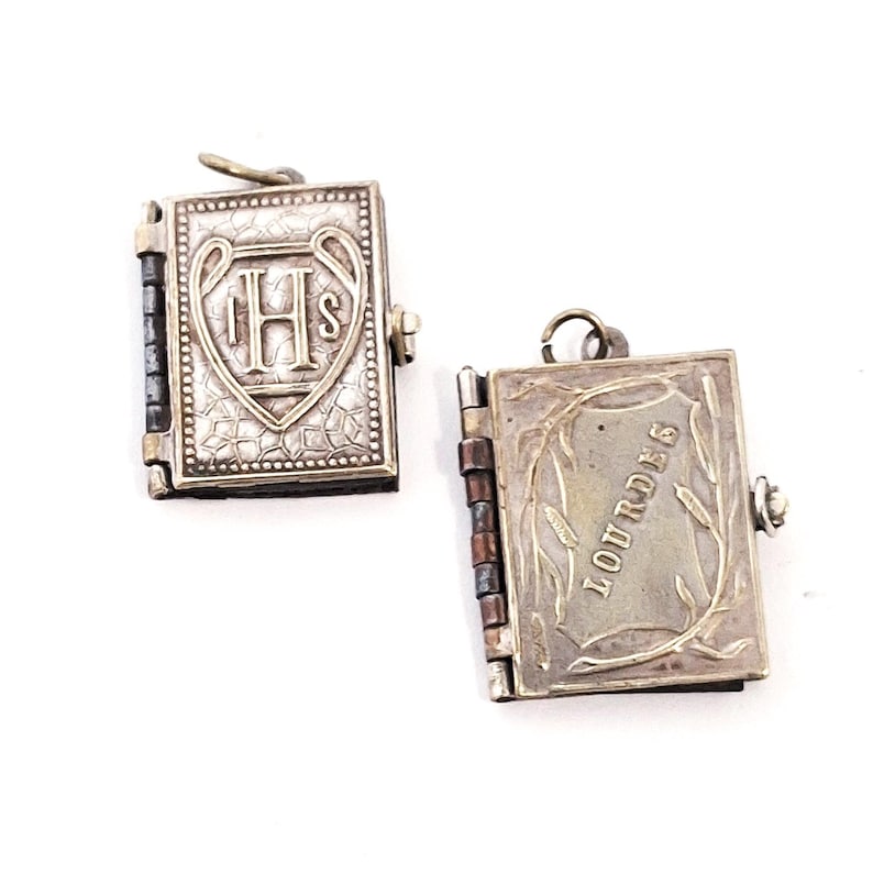May include: Two small, silver-coloured book-shaped pendants. One pendant features the letters "IHS" within a heart-shaped design. The other pendant is open, displaying the word "LOURDES" with a wheat stalk design. Both pendants have a hinged design.