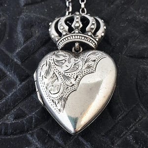 May include: A silver heart-shaped locket pendant suspended from a chain, featuring an ornate crown topper. The left side of the heart displays intricate engraved floral patterns, while the right side remains smooth and polished, reflecting light.