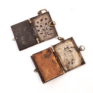 May include: Two open, antique silver lockets with hinged covers. One locket has a dark, textured interior and the other has a copper-coloured interior. The silver covers feature floral designs and small loops for hanging.