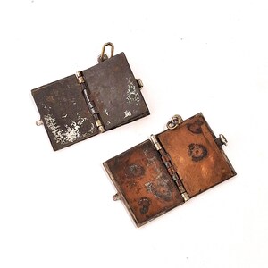 May include: Two small, open, rectangular pendants with hinged sides. The pendants are made of metal with a tarnished, aged appearance. One pendant has a dark, greyish-brown exterior and the other has a reddish-brown interior. Both have silver-coloured hinges and clasps.