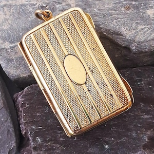 May include: A rectangular gold-toned metal locket featuring a textured cross-hatched pattern on the front surface, accented by vertical smooth stripes and a central blank oval cartouche. The locket includes a small circular bail at the top.
