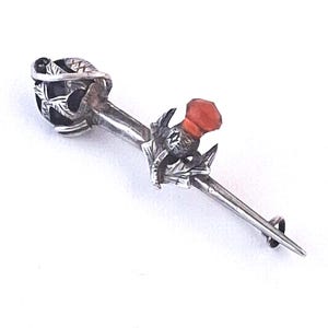 May include: A silver kilt pin featuring a thistle motif with a bright orange stone at the top and a dark, spherical stone encased in silver leaf-like metalwork at the opposite end, finished with a pointed tip and a small circular loop at the base.