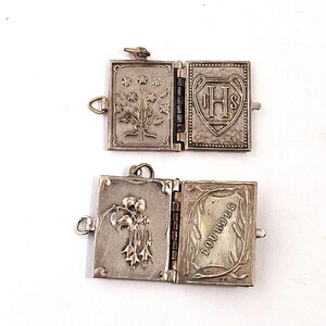 May include: Two open, silver lockets with hinged pages. One features floral designs and the letters IHS within a heart. The other has a floral design and the word "LOURDES".