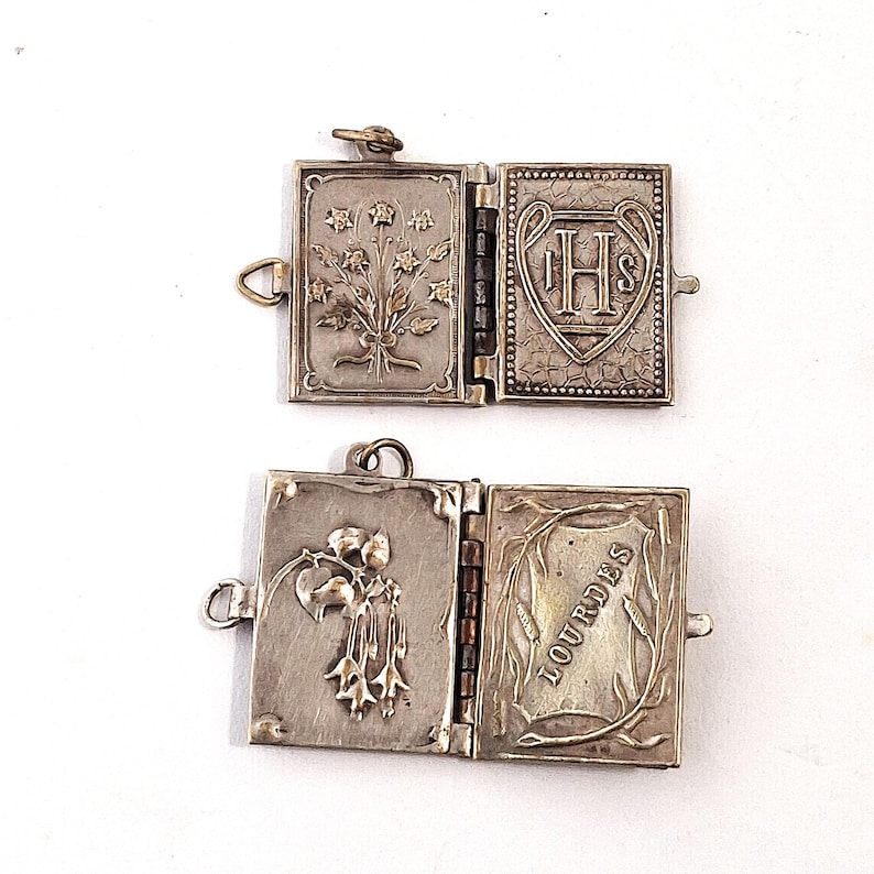 May include: Two open, silver lockets with hinged pages. One features floral designs and the letters IHS within a heart. The other has a floral design and the word "LOURDES".