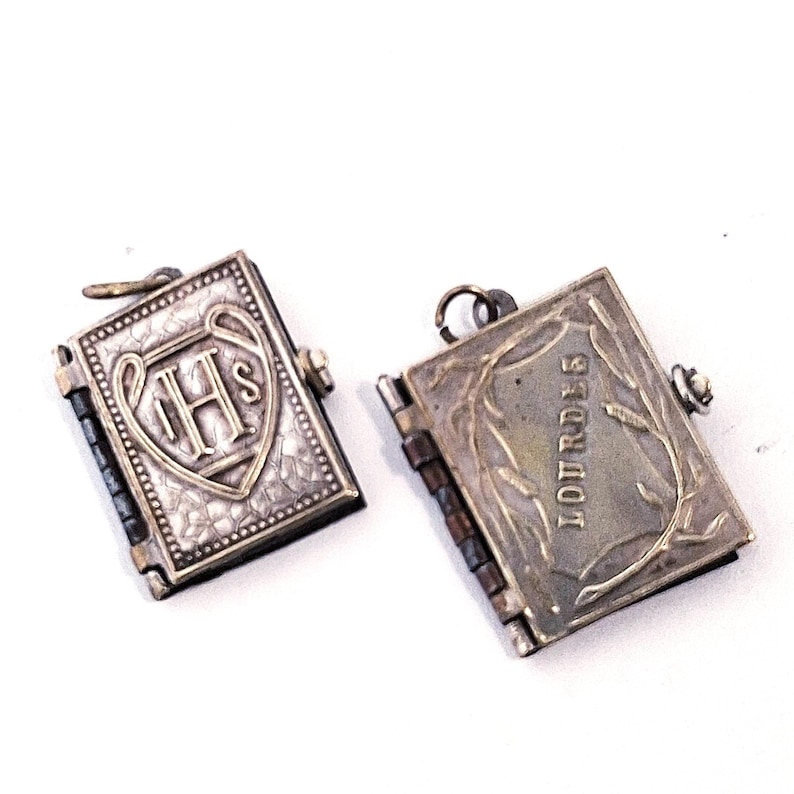 May include: Two small, silver-toned book-shaped pendants. One pendant features the letters "IHS" within a shield design. The other pendant is inscribed with the word "LOURDES" and a decorative border. Both have hinged spines.